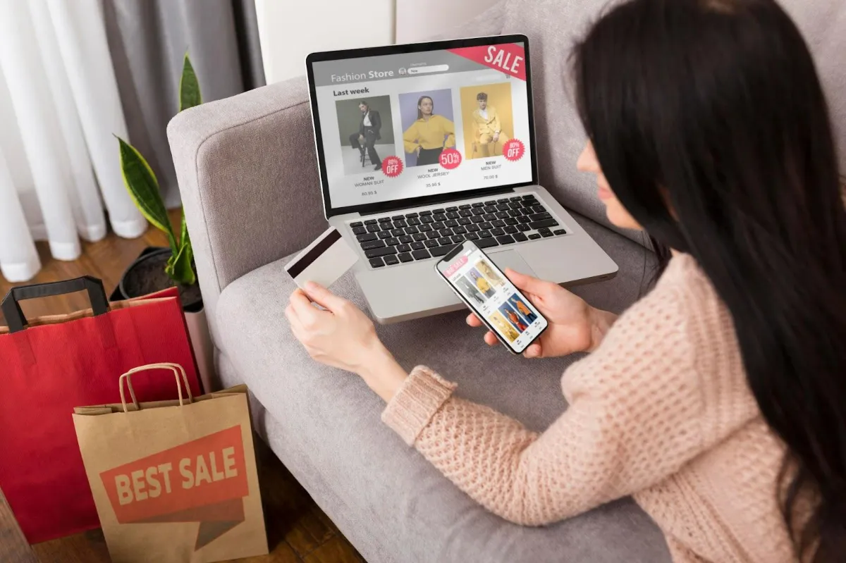 A person on a sofa shopping on a laptop and phone that both show sale promotions, with shopping bags beside them